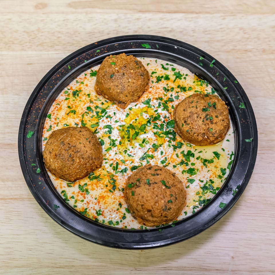 Four falafel balls sit in creamy tahini sauce, garnished with parsley, paprika, and olive oil in a black bowl.