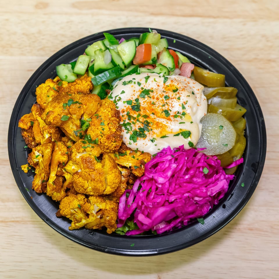 Black bowl of roasted spiced cauliflower, creamy sauce, cucumber salad, pickles, and bright purple cabbage slaw on a wooden table.