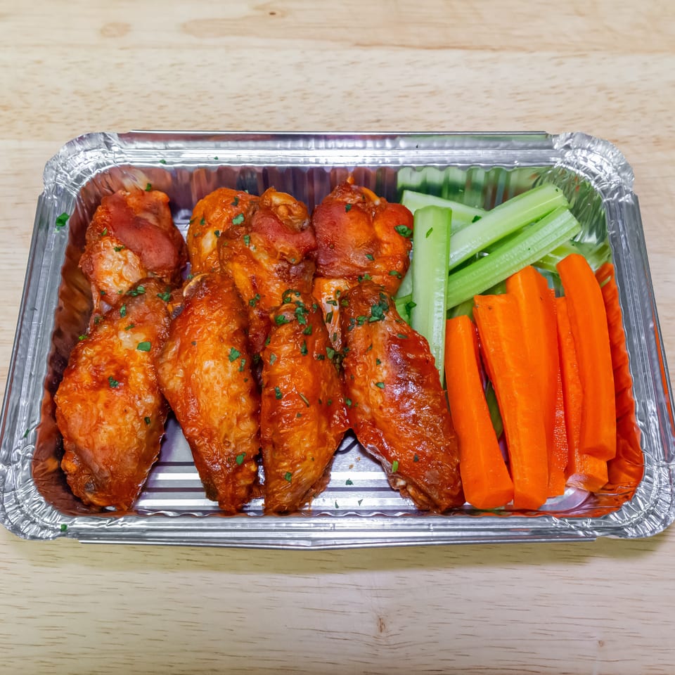 Aluminum tray of glazed chicken wings garnished with herbs, served with celery and carrot sticks on a wooden table.