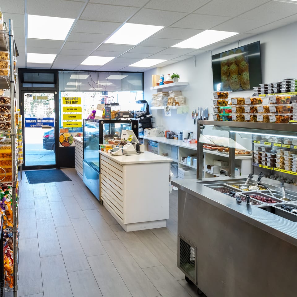 Bright deli shop interior with shelves of packaged snacks, refrigerated prepared foods, a service counter, and olive displays.