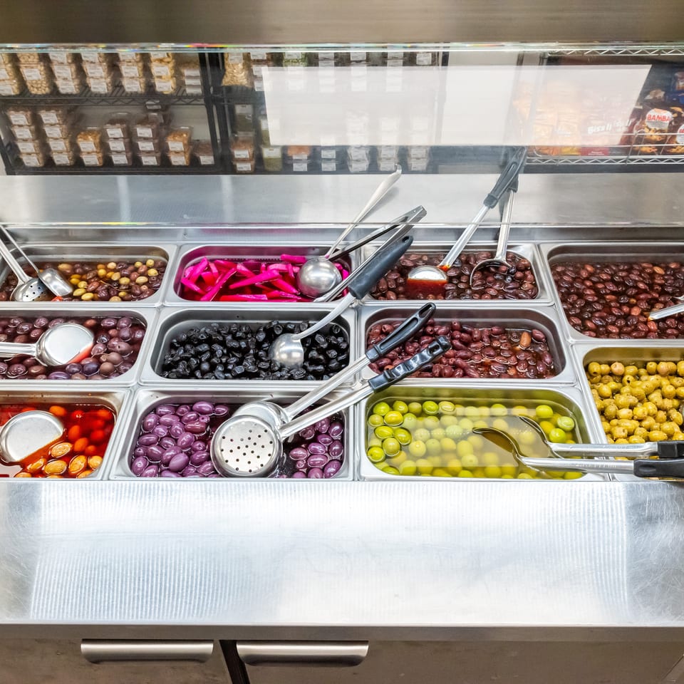 Stainless-steel counter displaying trays of assorted olives, pickled vegetables, serving spoons, and packaged foods on shelves.
