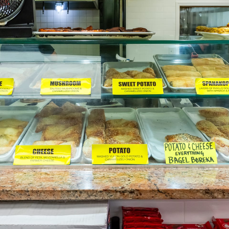 Bakery display case filled with labeled vegetable, mushroom, sweet potato, spanakopita, potato, cheese, and spinach pastries.