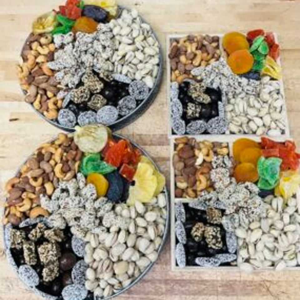 Four serving trays filled with assorted nuts, dried fruits, colorful candied fruits, and powdered treats on a wooden table.